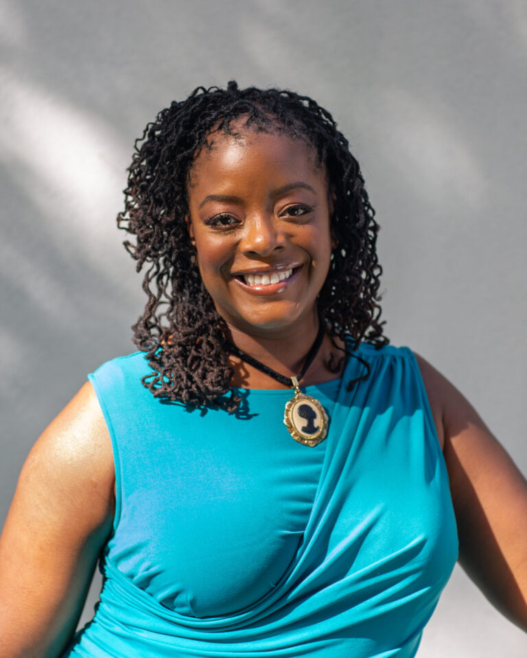 A Black woman with curly locs, smiling, wearing an aqua dress with a Black cameo necklace.