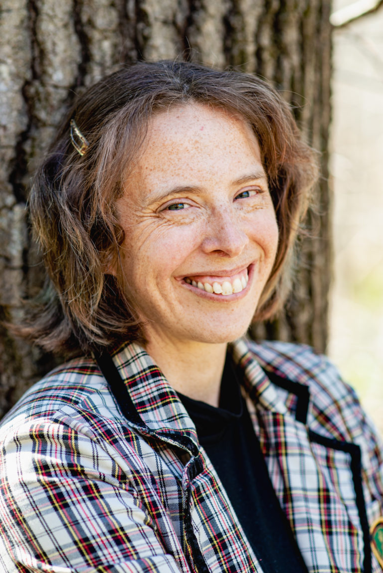 A white woman in her 40s with brown hair, a plaid coat, and a black shirt leans against a tree smiling.