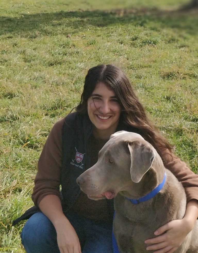 A Mexican-American woman with long brown hair and brown eyes, wearing a brown sweater and black vest, with a gray dog in front of her. 