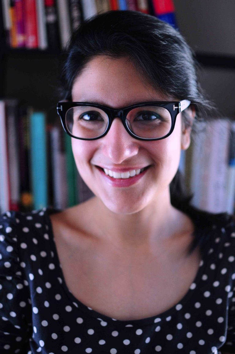 A South Asian woman with long black hair pulled in a pony tai lwearing thick-rimmed black glasses and a black blouse with white polka dots smiling with bookshelves in the background.