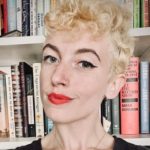 White woman with a blonde curly pixie cut wearing a black shirt standing in front of a full bookshelf (shoulders up)