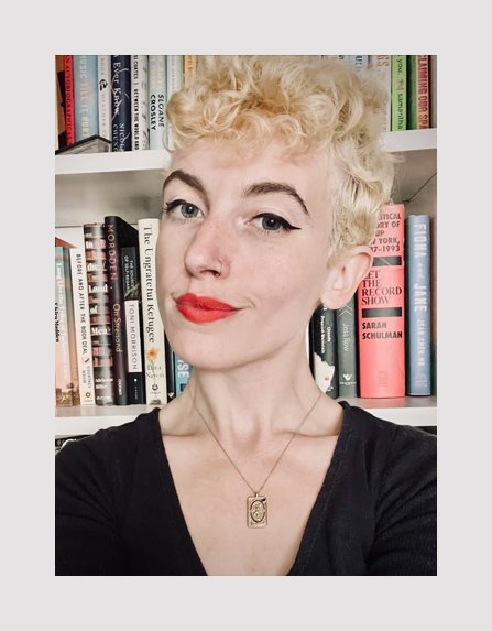 White woman with a blonde curly pixie cut wearing a black shirt standing in front of a full bookshelf (shoulders up)