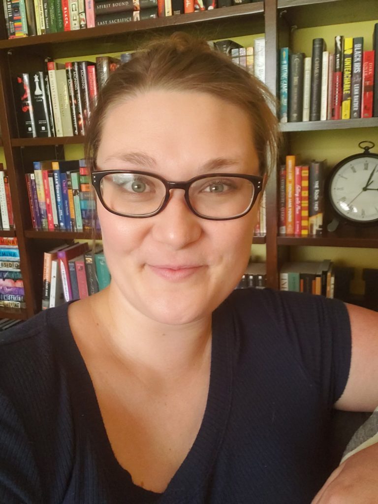 A white cis woman in her late 30s with dark blonde hair, dark-rimmed glasses, and a wall of books behind her.