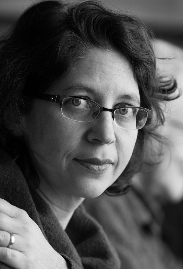 A mixed-race (white/South Asian) woman in her forties with glasses and mid-length greying brown hair.