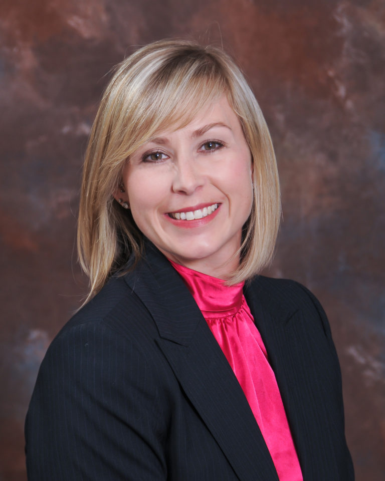 Ann Rose is a cis, white woman. In the photo she has blonde hair that has been cut into a bob, with sweeping fringe. She is of average build, and is wearing a suit jacket with hot pink shirt underneath. She is set in front of a natural background.