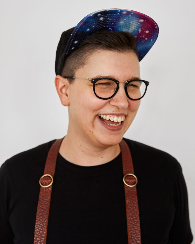 A white nonbinary person, smiling big and wearing a ball cap with a galaxy-print brim. They are clearly having a rad time.