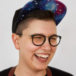 A white nonbinary person, smiling big and wearing a ball cap with a galaxy-print brim. They are clearly having a rad time.