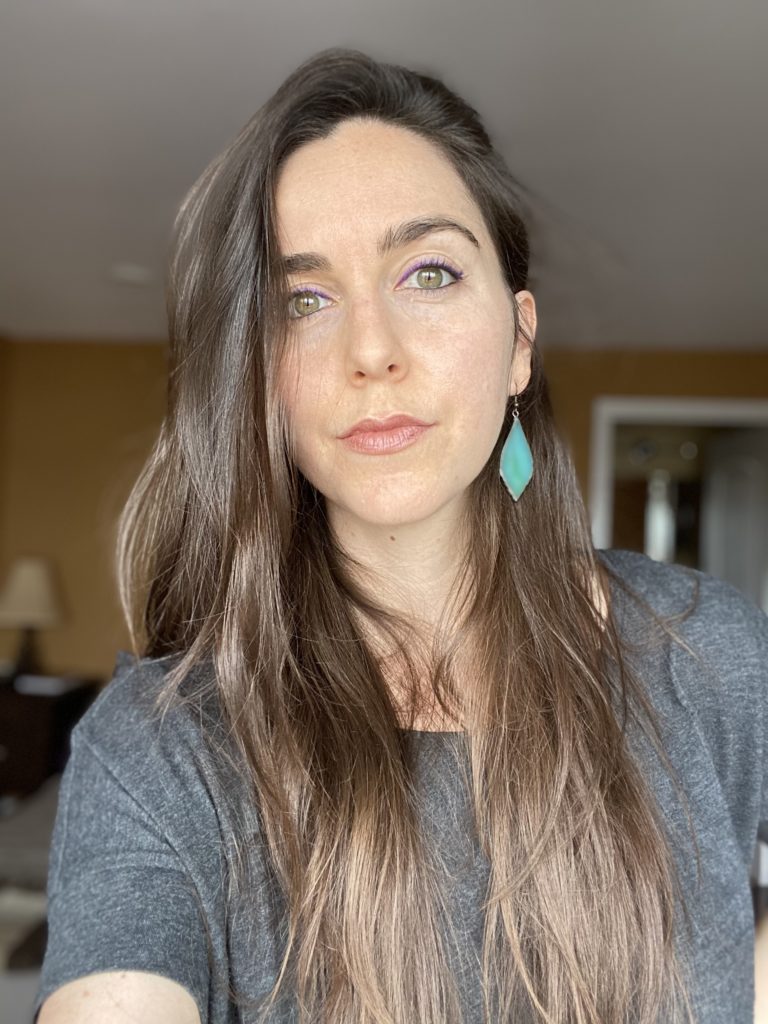A white woman in her early thirties, with teal earrings, long brown hair, and a gray shirt.
