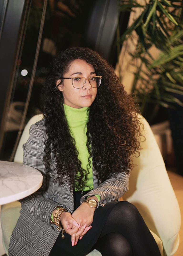 Paloma seated at a chair. She wears a neon green turtleneck and a grey plaid blazer. She smiles slightly. she wears glasses and has curly brown hair.