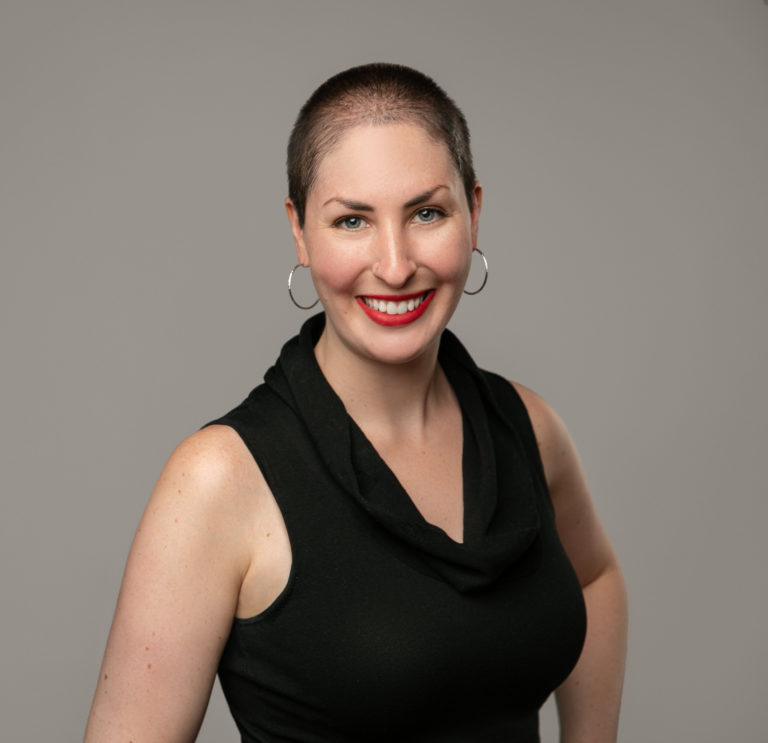 A white woman in her early 30s, with a buzz cut, hoop earrings, and a black top.