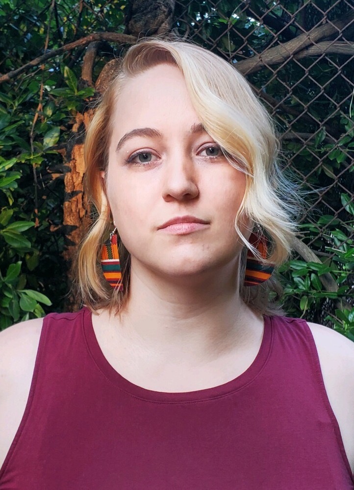 A white nonbinary person in their thirties, with shoulder-length wavy blonde hair, wearing burgundy.