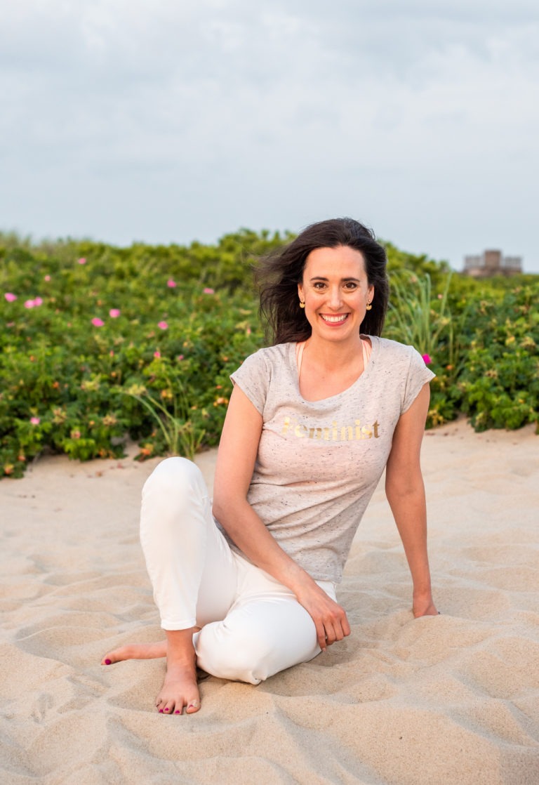 A tall, dark-haired white woman wearing a t-shirt that says "Feminist" sits on a beach, smiling at the camera.