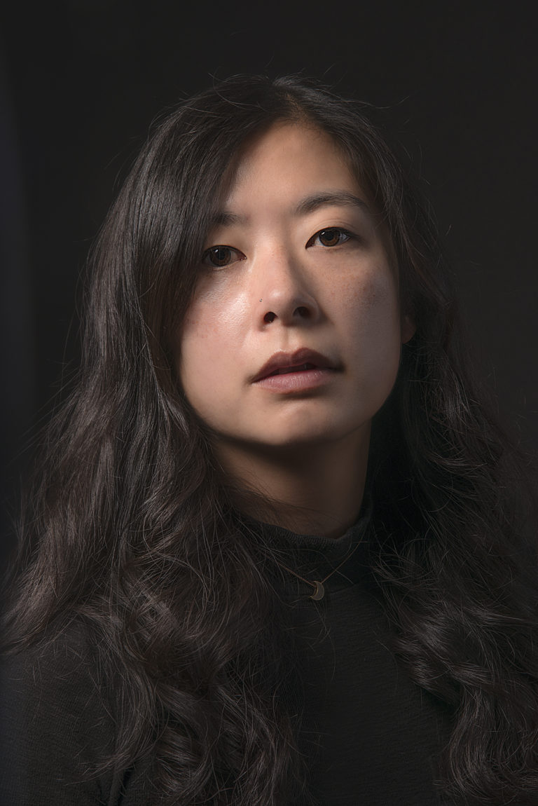 An Asian-American cisgender woman with long hair in front of a black background.