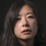 An Asian-American cisgender woman with long hair in front of a black background.