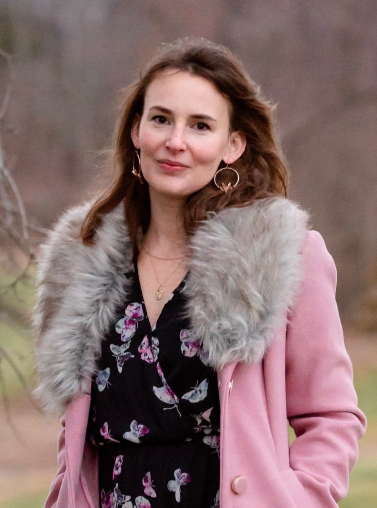 A white woman in her thirties, with shoulder-length brown hair, wearing hoop earrings and a pink coat with a fake fur collar.