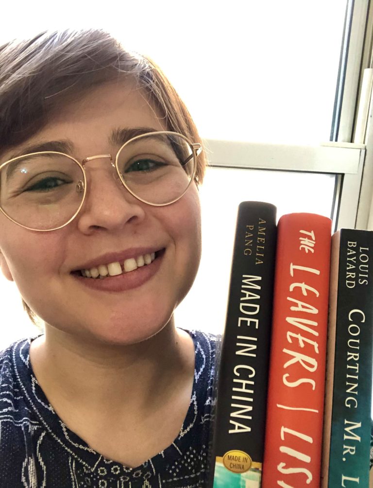 A white woman in her late twenties, with short straight brown hair and wire-rimmed roundish glasses. She is holding up several books and smiling.