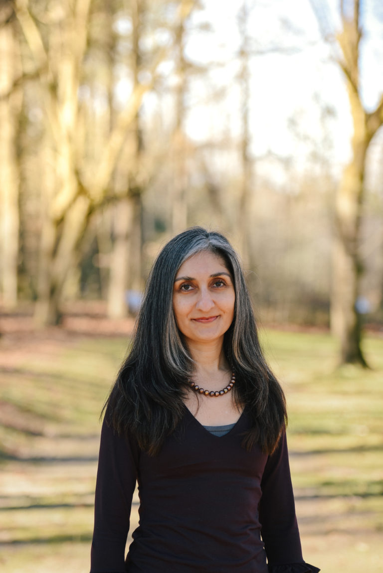 Brown-skinned woman in late forties with dark brown and white layered straight hair that falls past shoulders. Brown long sleeve v-neck shirt, brown pearls. In the background, a woods with bare trees.