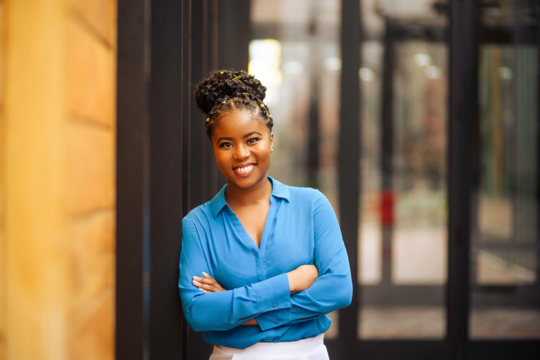 A brownskin Black woman in her 20s with her arms crossed and black twists in a bun wearing a blue blouse and white pants. She can be seen from the waist up and is smiling at the camera.