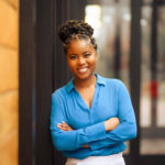 A brownskin Black woman in her 20s with her arms crossed and black twists in a bun wearing a blue blouse and white pants. She can be seen from the waist up and is smiling at the camera.