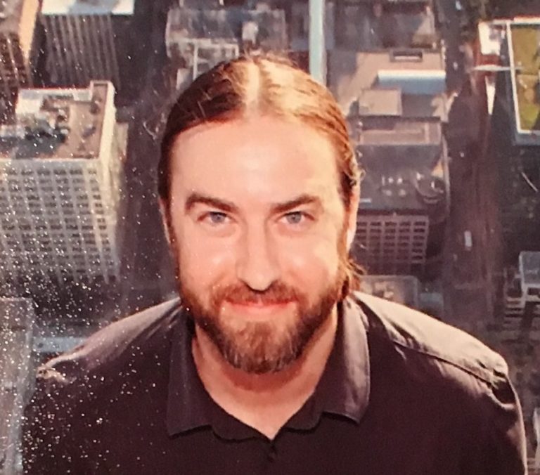 A bearded white man in a black shirt in front of a cityscape