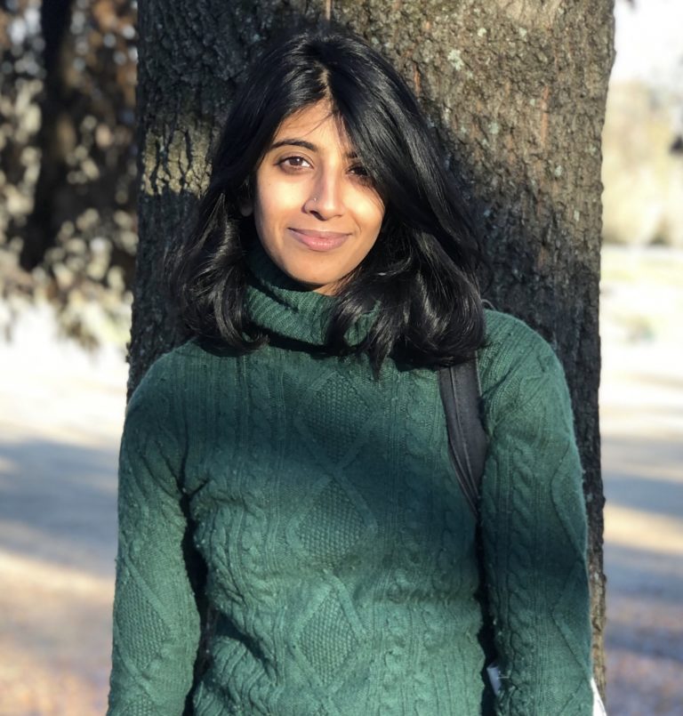 Indian American cisgender woman, black hair, green sweater, leaning against a tree.