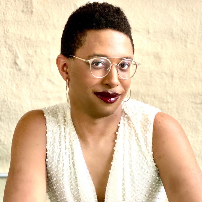 A Black trans writer and editor in her mid thirties, who looks in her mid twenties, wearing a tank top and a dark red lipstick. Her hair is a cropped short natural.
