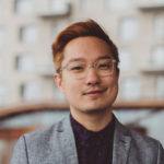 A portrait of queer Chinese American poet Chen Chen, standing in front of a large beige apartment building. His hair is dyed an orangey-blonde. He wears clear-framed glasses, a dark purple button-down with gray polka dot pattern, and a gray blazer. His facial expression is gently joyful.
