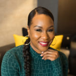 African American woman in her forties wearing a long-sleeve teal knit dress and hair pulled back in a ponytail with a side part, sitting on a leather sofa with hand on chin bearing a big smile.