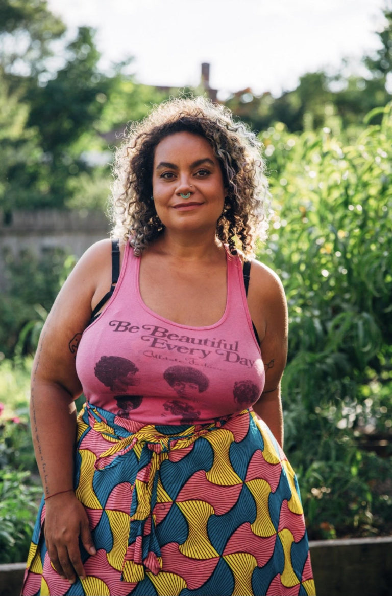 Black mixed race woman with curly hair in pink tank top that says Black is Beautiful and a Kenyan skirt in pink and yellow, outdoors in Detroit nature.