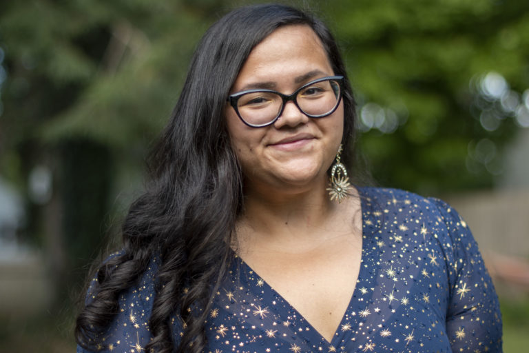 A fat, Puerto Rican woman in her mid-thirties with long, dark, curly hair, black cateye glasses, and wearing a small smile and a star-speckled navy and gold dress.