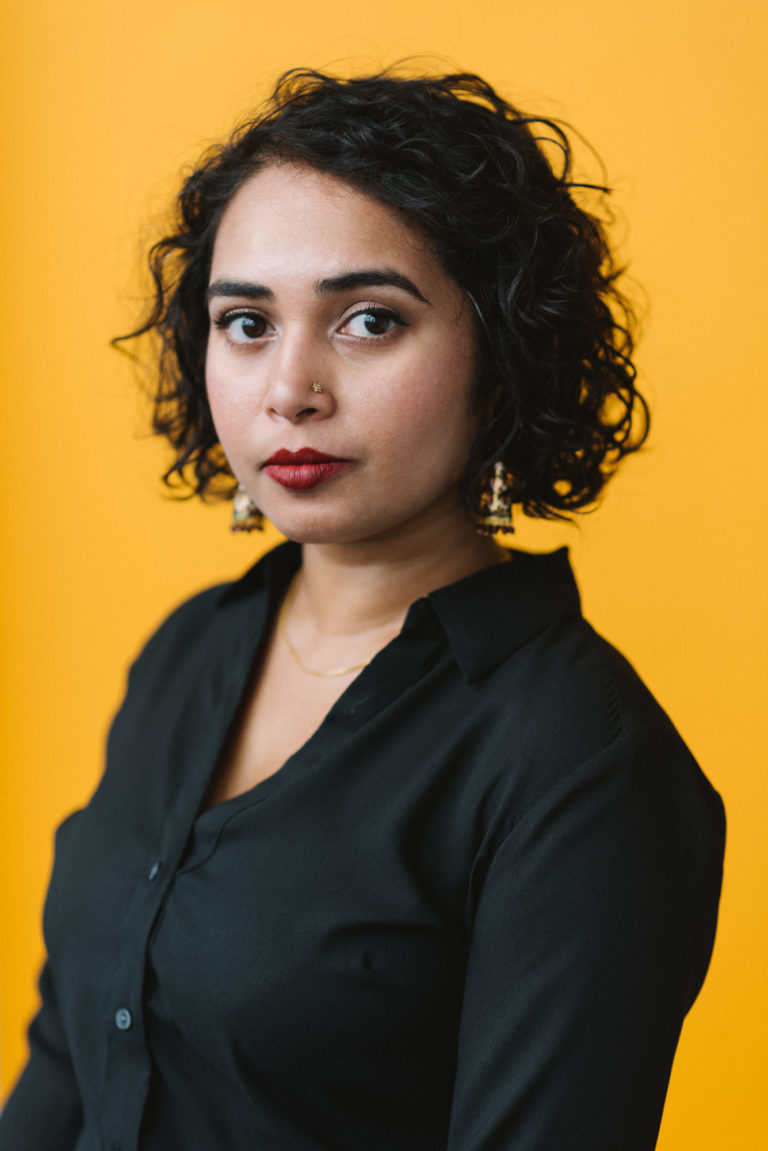 A light-skinned South Asian queer femme with short curly hair and red lipstick, in her early-thirties, wearing a black blouse.