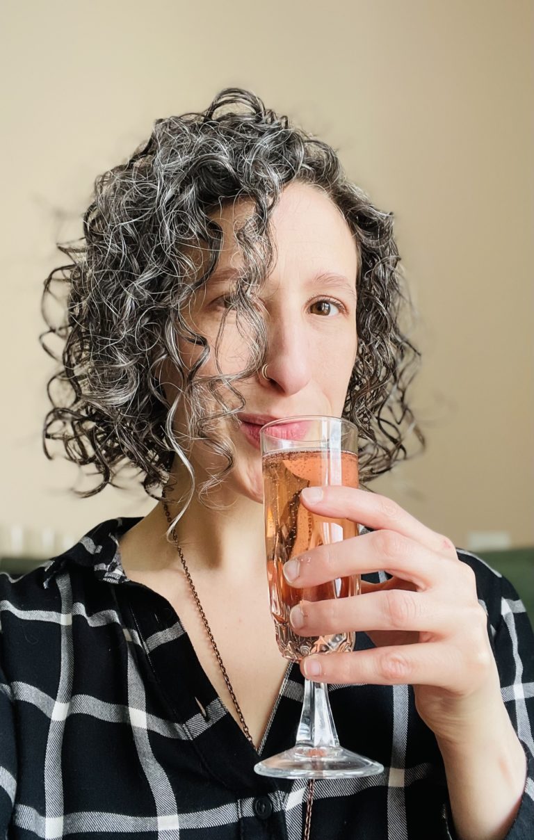 A white woman in her late 30's with short dark curly hair and a black top regards the camera while holding a champagne flute filled with pink liquid to her lips.