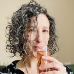A white woman in her late 30's with short dark curly hair and a black top regards the camera while holding a champagne flute filled with pink liquid to her lips.