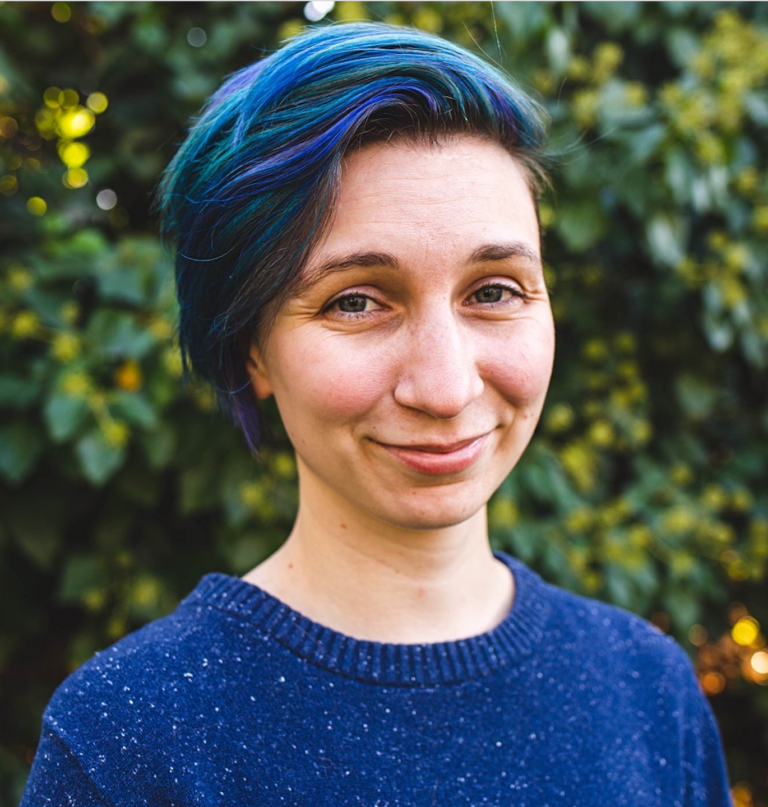 A nonbinary white person in their late 20s, with short blue hair and wearing a blue sweater.