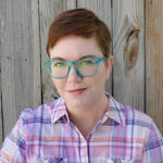 A nonbinary white person with short red hair, blue eyes, and teal glasses. They're wearing a pink plaid collared shirt in front of a wooden background.