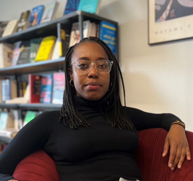 A Black woman in her twenties with braids, clear chunky-framed glasses, a black turtleneck and very light blue jeans. She is sitting in a plush cranberry colored chair with her left arm draped on its arm.