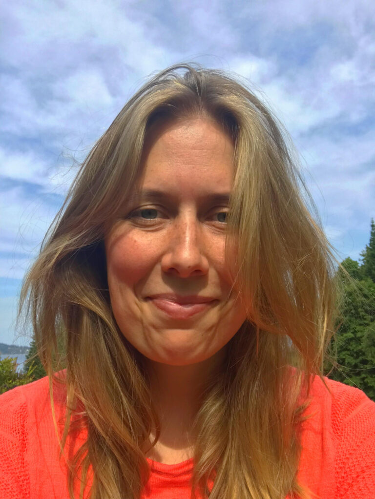 A blond woman in her 30s in a coral shirt, outside.